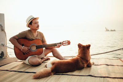 Man playing guitar while sitting on pier by dog against sky
