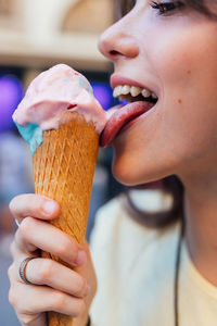 Crop smiling young female licking tasty gelato in waffle cone while looking away in town on blurred background
