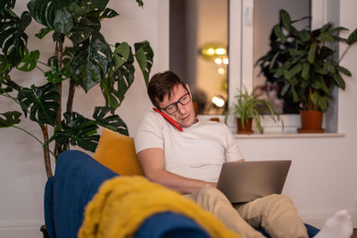 Busy man sitting on couch with laptop. serious guy with glasses talking on phone, focused on work