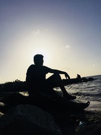 Man sitting on rock at beach against sky during sunset