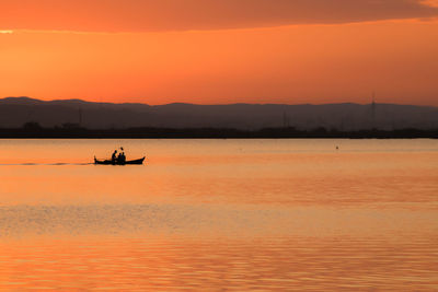 Silhouette men on boat in lake against orange sky