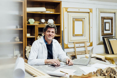 Smiling craftsman sitting with tablet pc at workbench in workshop