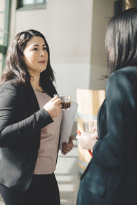 Female professionals having coffee while discussing in office corridor