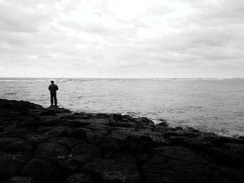 Rear view of young man fishing while standing at beach against cloudy sky