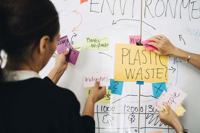 Rear view of businesswoman with colleague arranging adhesive notes on whiteboard at creative office