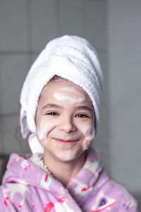 A girl with a white bath towel on her head and cleansing foam applied to her face smiles