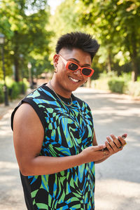 Happy filipino transgender woman in stylish clothes and sunglasses browsing cellphone and looking at camera with smile while standing on sunlit path in summer in park