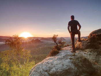 Man on the rock empire with hands in trousers pocket watch over misty valley to sun at horizon.