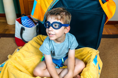 Boy wearing eyewear looking away while sitting in open suitcase