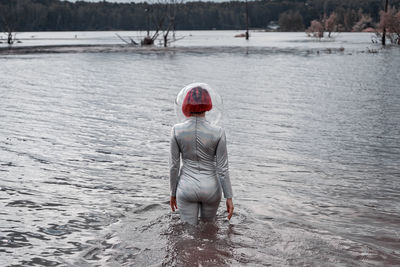 Back view of unrecognizable futuristic young red haired female in silver space suit and glass helmet walking on flooding river