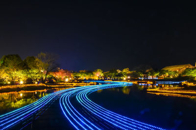 Light trails on river by illuminated city against sky at night