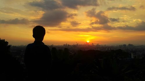 Silhouette boy standing by cityscape against sky during sunset