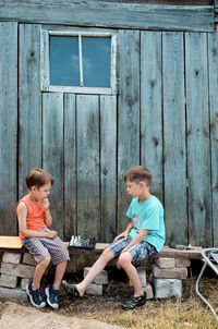 Two village boys play chess on the street, sitting on a bench, against the backdrop of an old barn