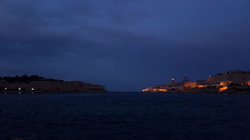 Illuminated buildings by sea against sky at dusk