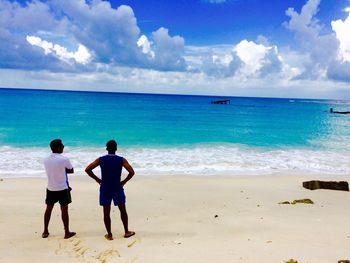 Rear view of friends standing on sea shore at beach against blue sky