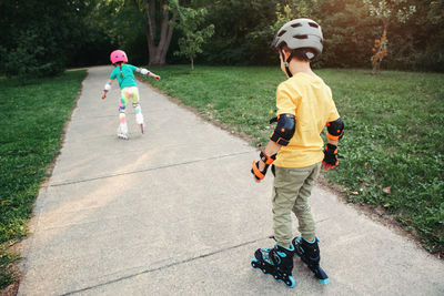 Friends boy and girl in helmets riding on roller skates in park on summer day. 