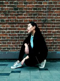 Young woman looking away while sitting against brick wall