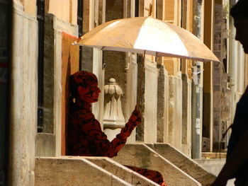 Side view of young woman with umbrella sitting on steps by building in city