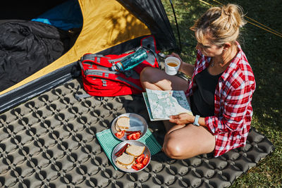 Woman having breakfast and planning day sitting by tent at camping. female spending vacations