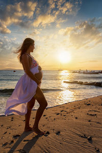 Pregnant woman walking at beach against sky during sunset