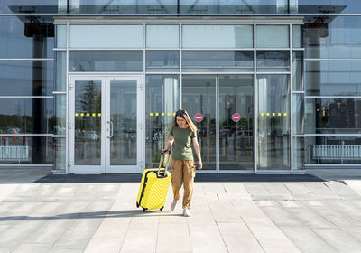 Young woman traveler in casual carrying yellow suitcase next to entrance to airport outside tourism