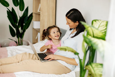 A young mother is working on a laptop or watching videos with her little daughter in the bedroom. 