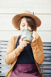 Woman having coffee while sitting on wooden bench against wall