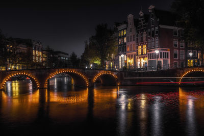 Illuminated arch bridge over river by buildings in city at night