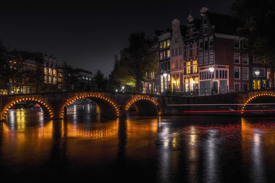 Illuminated bridge over river by buildings in city at night