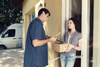 Delivery man giving pen and clipboard to woman against house