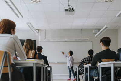 Students sitting at desk while teacher writing on whiteboard in classroom