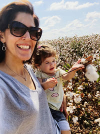 Portrait of smiling woman wearing sunglasses standing with daughter at farm against sky