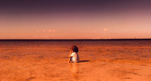 Rear view of baby girl sitting at beach during sunset