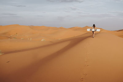 Back view of young female walking on sand and preparing for sandboarding looking away