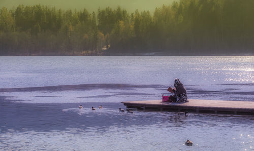 Woman sitting on jetty over lake during sunny day against trees