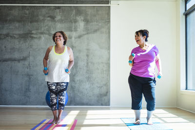 Full length of happy female friends using dumbbells while exercising against wall in yoga studio