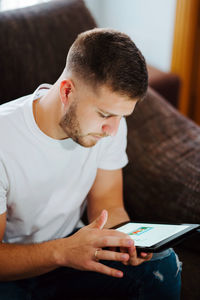 Delighted young male relaxing on couch and surfing internet on tablet while enjoying weekend at home