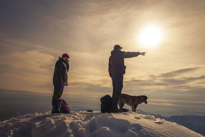 People with dog on the snow summit against sky during sunset