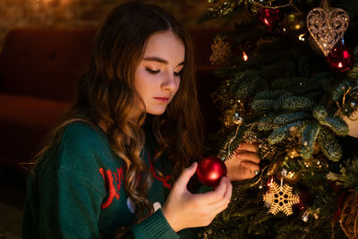 Cute teenager girl decorates christmas tree with red balls in green sweater