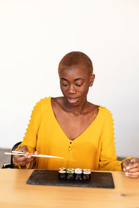 Young woman with sushi on table sitting against white wall at home