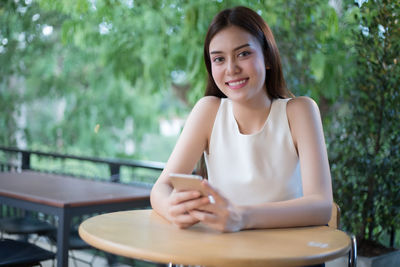 Portrait of smiling young woman using phone while sitting at sidewalk cafe