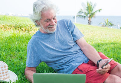 Senior man using mobile phone and laptop while sitting on grassy land