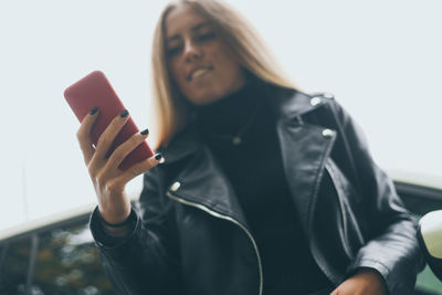 Trendy blonde girl using smartphone leaning against her car in a beautiful autumn afternoon. 