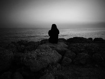 Silhouette woman sitting on rock by sea against clear sky