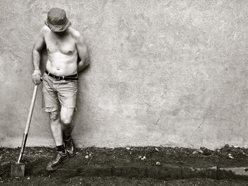 Full length of shirtless man standing with shovel against wall