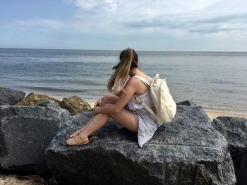 Rear view of woman sitting on rock by sea against sky