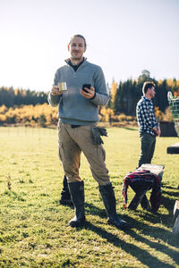 Full length portrait of confident farmer holding coffee cup and mobile phone on field