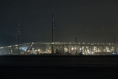 Illuminated bridge over river by buildings against sky at night