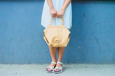 Low section of woman holding handbag while standing in front of blue wall