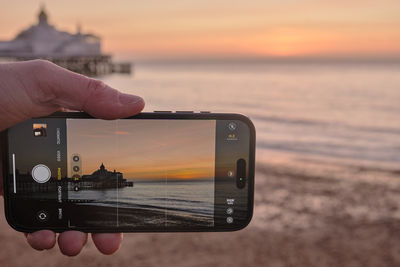 Cropped hand of man photographing sea against sky during sunset